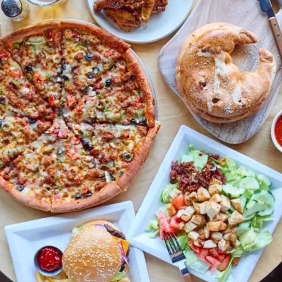 Overhead view of a restaurant spread featuring topped pizza, stuffed bread, grilled chicken salad, and a sesame-seed burger.
