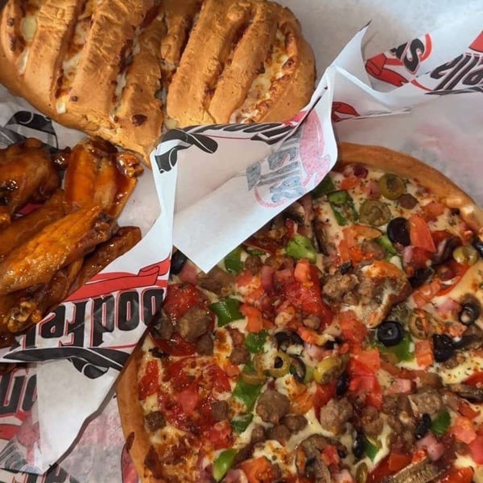 Loaded pizza with vegetables, olives, meat, glazed chicken wings, and toasted bread served on branded paper.